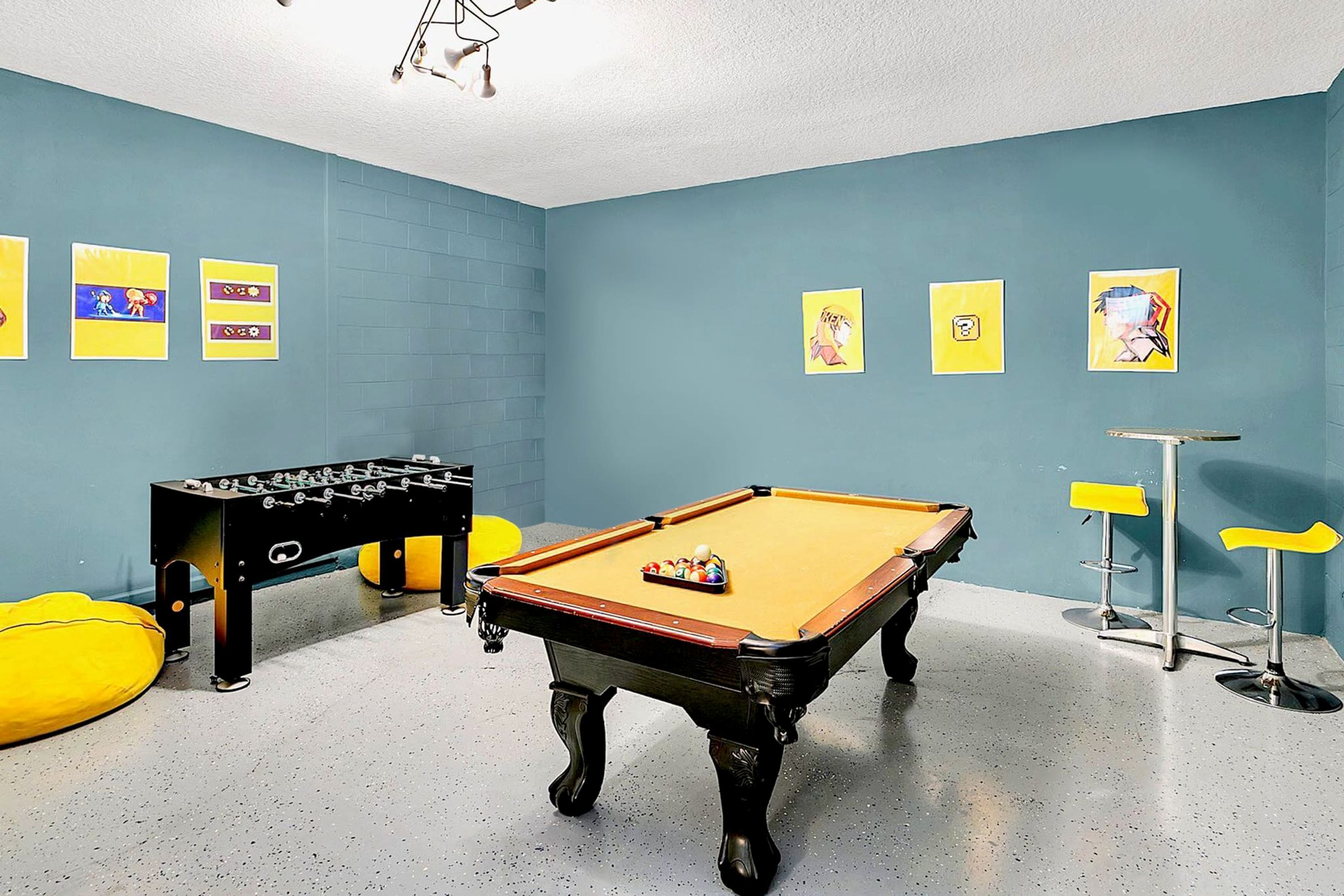 A pool table with a foosball table beside it, a bar area with stools, and several framed pictures on the walls.