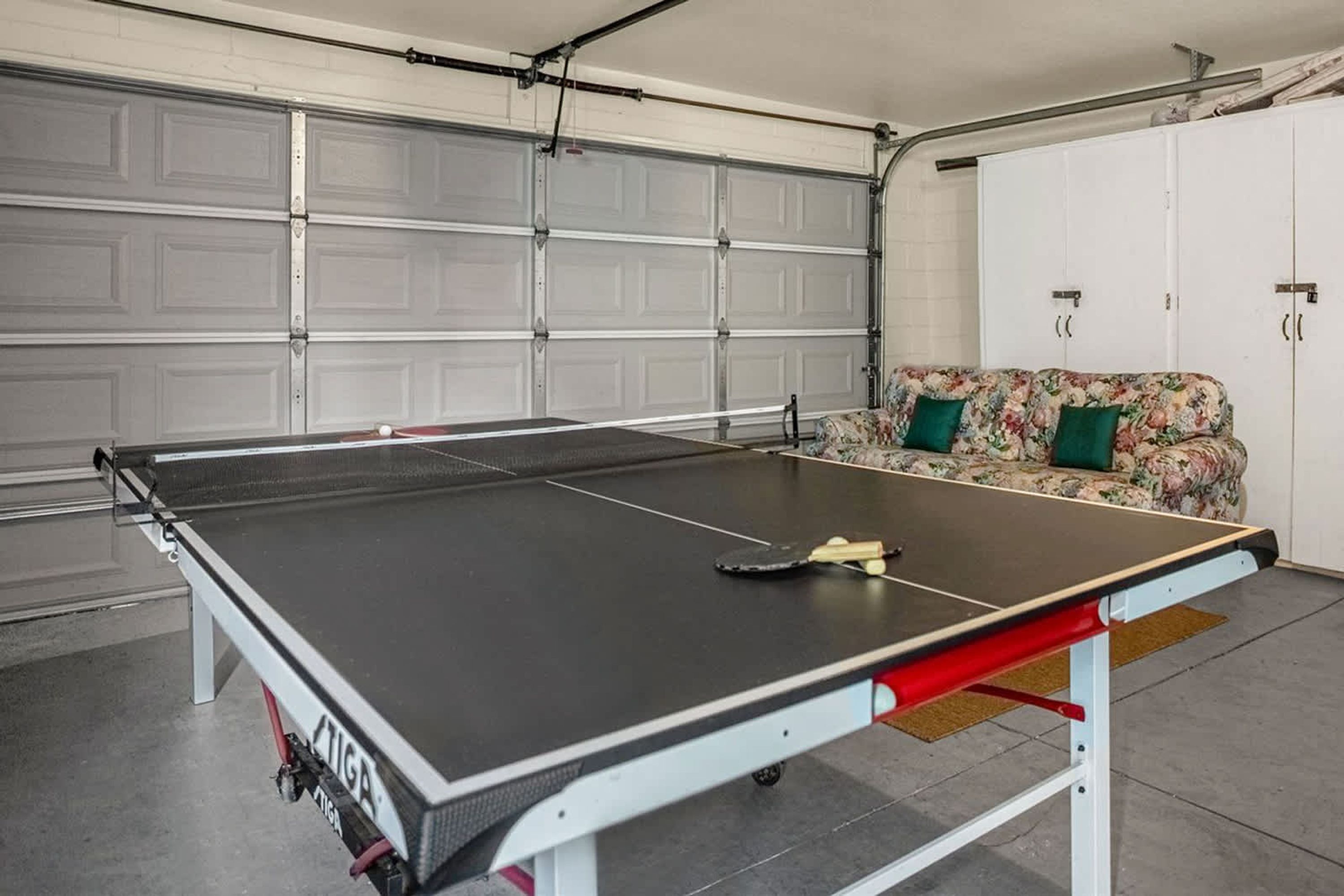 Une table de ping-pong dans un garage, avec un canapé à motifs floraux et des armoires.