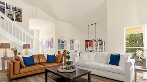 Ein geräumiges Wohnzimmer mit einem braunen Ledersofa, einem weißen Sofa und einem dunklen Couchtisch. Eine Treppe mit weißen Geländern und ein großes Fenster mit Blick auf Grünpflanzen.