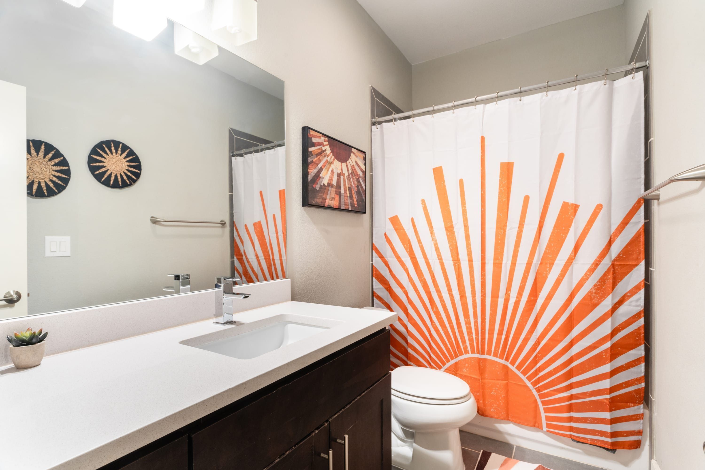 Un baño con un lavamanos blanco, muebles oscuros, un inodoro blanco y una cortina de ducha con un diseño de sol naranja.