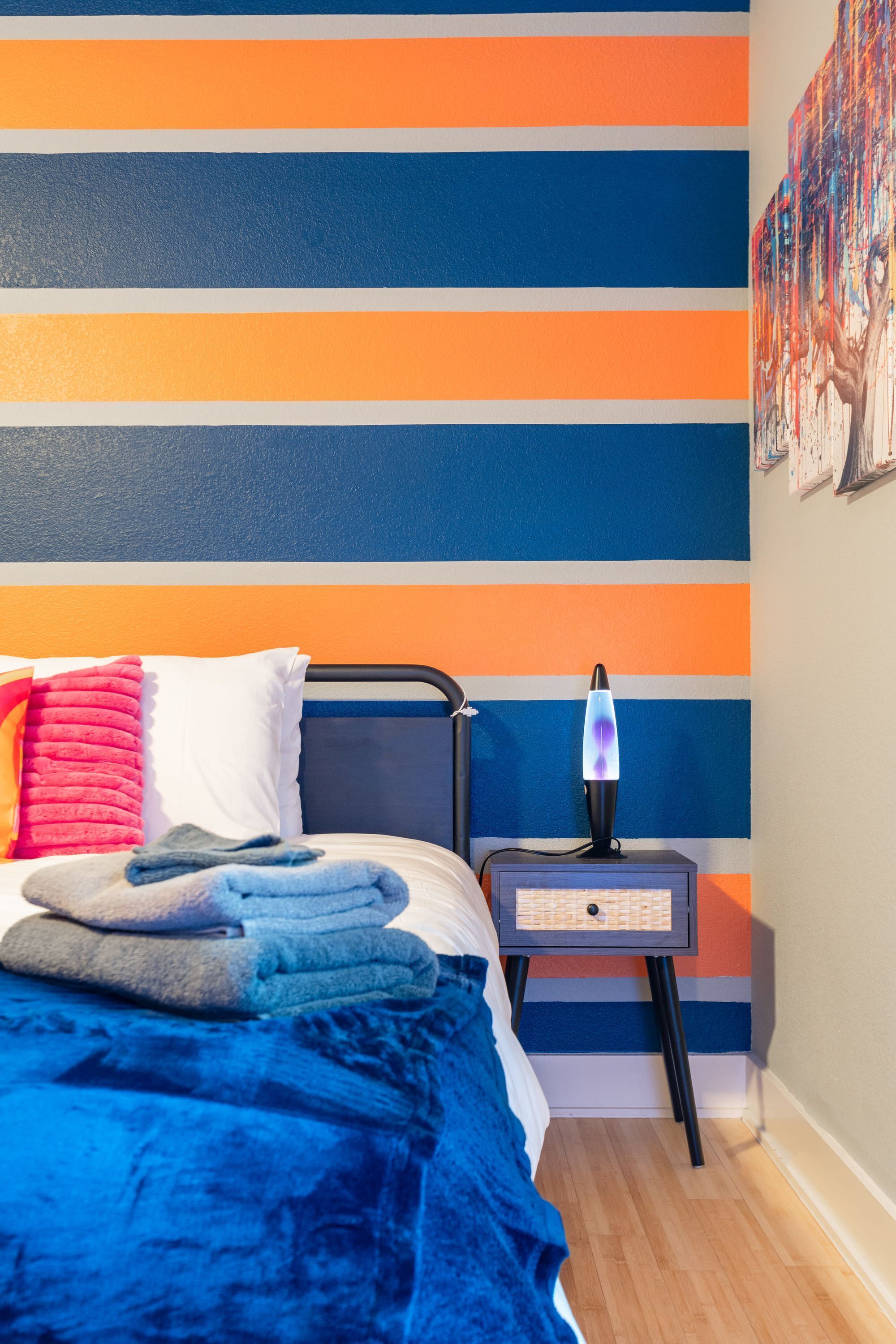 Un dormitorio con cama, almohadas, una colcha, una mesita de noche con una lámpara de lava y una pared con rayas horizontales.