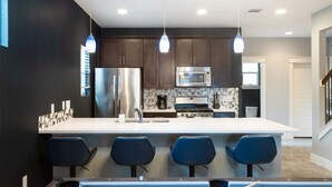 A modern kitchen with dark cabinets, a stainless steel refrigerator, a microwave, and a coffee maker. There are four blue bar stools around a central island. - Upstay - East Austin Home w Yard Games Fire Pit (Austin)