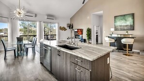 A modern kitchen with a large island, a dining area with a glass table, and a bar area with stools. - Upstay - Lakeside Getaway - Pool Hot Tub Games (Lakeway)