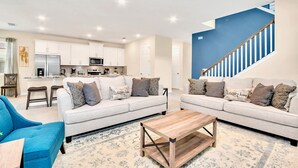 Un salon moderne avec deux canapés, une table basse et un fauteuil bleu. On aperçoit, au fond, une cuisine avec des meubles blancs et des appareils électroménagers.