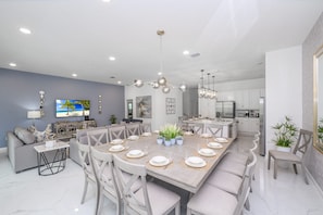 A modern kitchen with white cabinets, a dining area with a large table set for six, and a living area with a sofa and a TV. - Luxury Private Pool 26 Guest Capacity Mansion (Kissimmee)