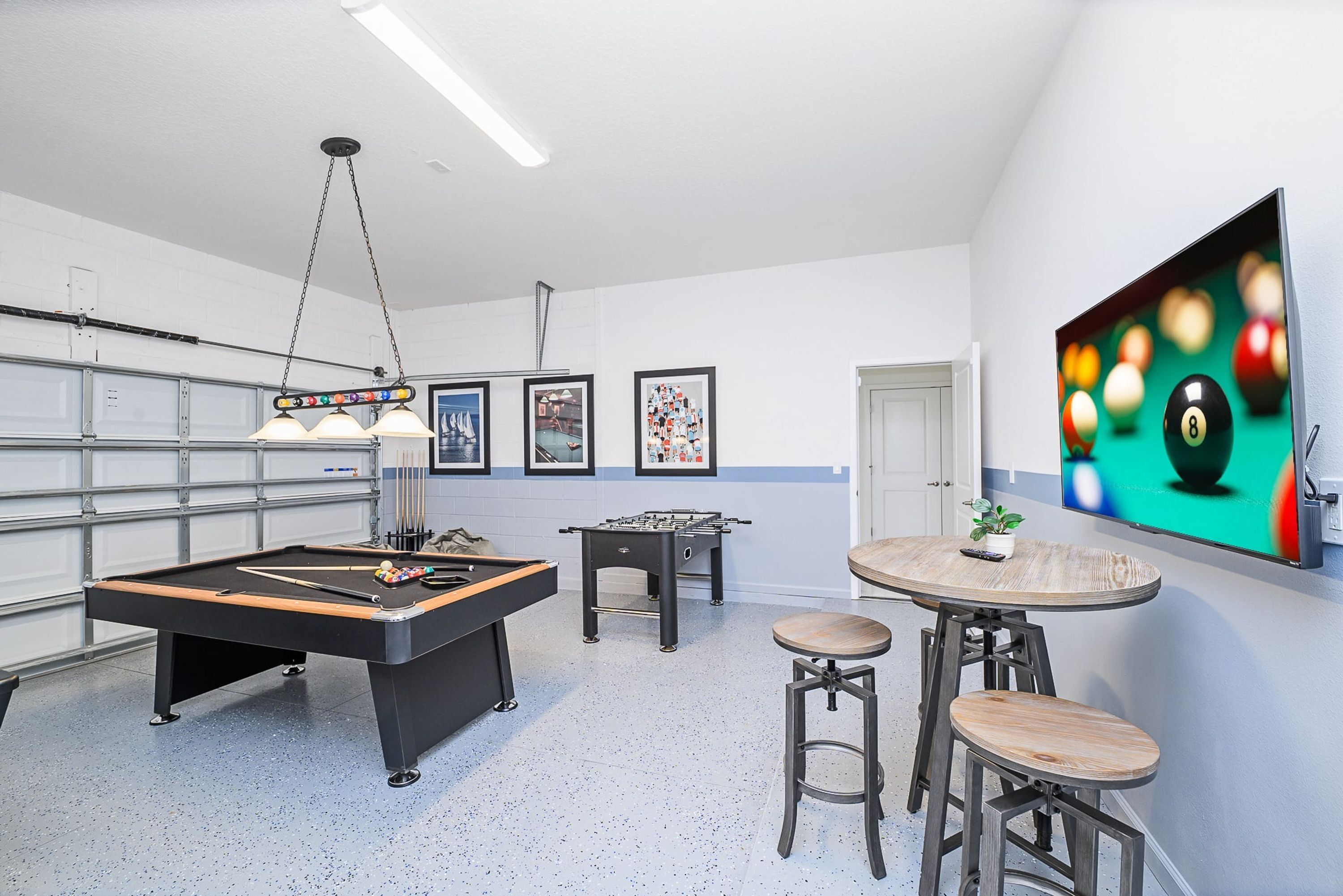 A pool table, foosball table, and bar stools in a room with a large TV screen and framed pictures on the wall.