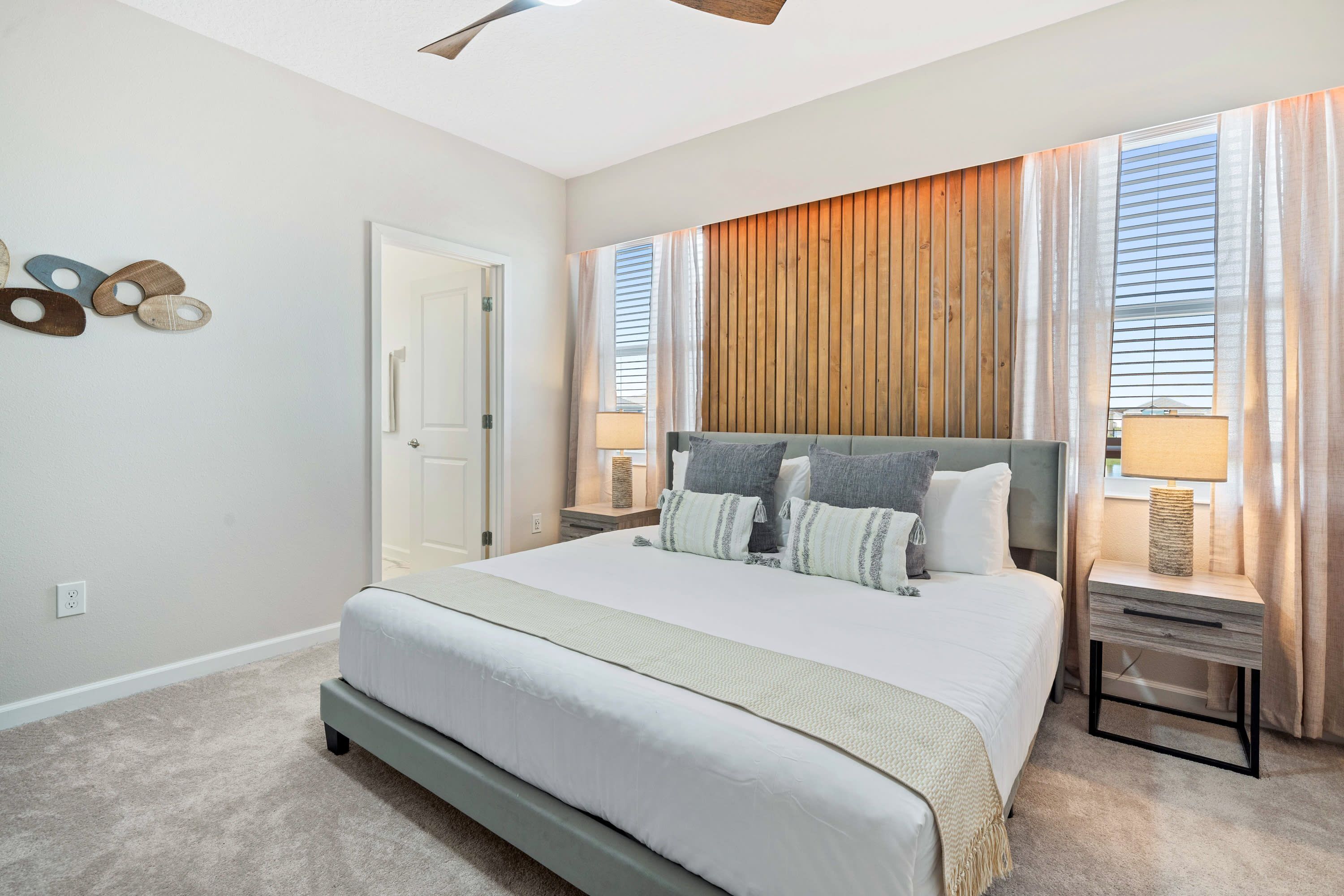 A bedroom with a large bed, a ceiling fan, a wooden panel wall, and a window with curtains.