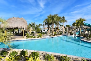 A swimming pool with a water slide, surrounded by palm trees and a thatched-roof structure.