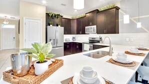 Una cocina moderna con armarios oscuros, una encimera blanca y una isla central. Hay plantas en macetas sobre los armarios y un lavamanos de acero inoxidable.