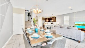 Un área de comedor moderna con una mesa larga dispuesta para seis personas, un sofá gris y una cocina al fondo.