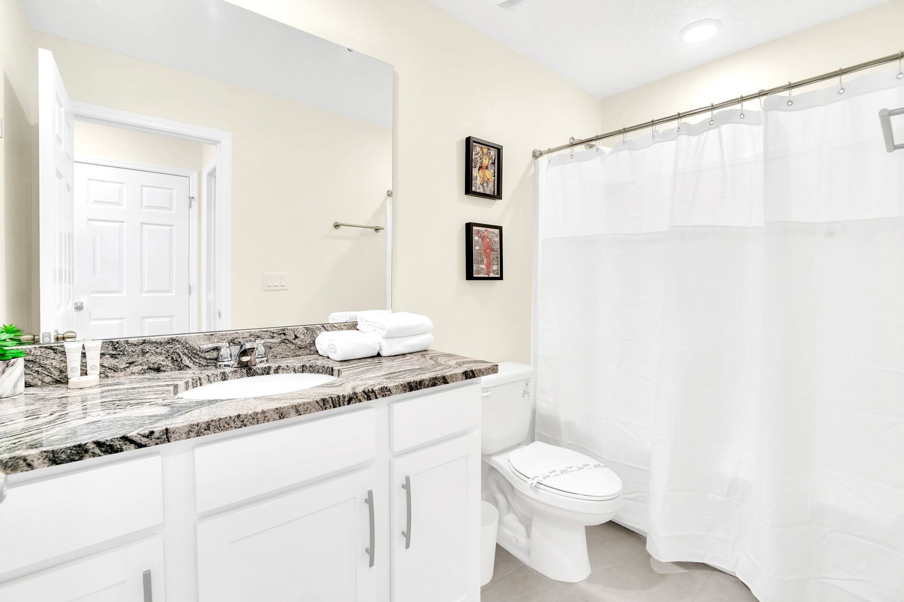 A bathroom with a white sink, marble countertop, white cabinets, a white toilet, and a white shower curtain.