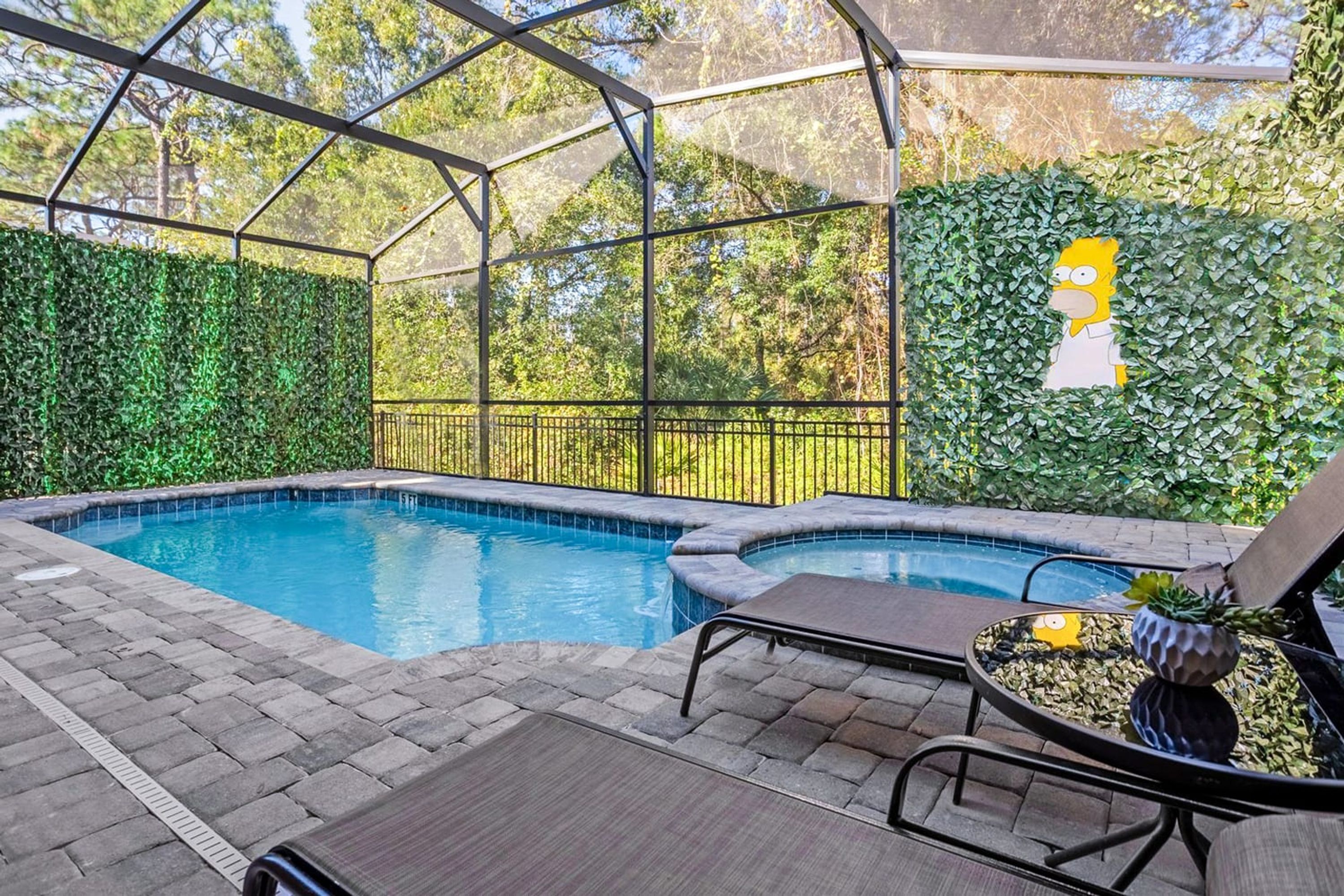 A covered outdoor swimming pool with a glass enclosure, surrounded by a hedge and a metal fence.