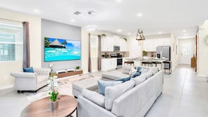A modern living room with a large sectional sofa, a flat-screen TV, and a dining area with a kitchen in the background. - Upstay - 20-guest Home w Hot Tub-pool-bbq-arcade (Kissimmee)