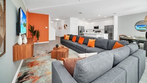 A modern living room with a grey sectional sofa, orange cushions, and a wooden coffee table. - Upstay - Modern Pool Home w Games - Sleeps 18 (Kissimmee)