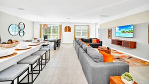 A modern living room with a grey sofa, a dining area with a long table, and a TV mounted on the wall. - Upstay - Modern Pool Home w Games - Sleeps 18 (Kissimmee)