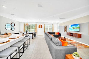 Ein modernes Wohnzimmer mit einer grauen Couch, einem Essbereich mit einem langen Tisch und einem an der Wand befestigten Fernseher.