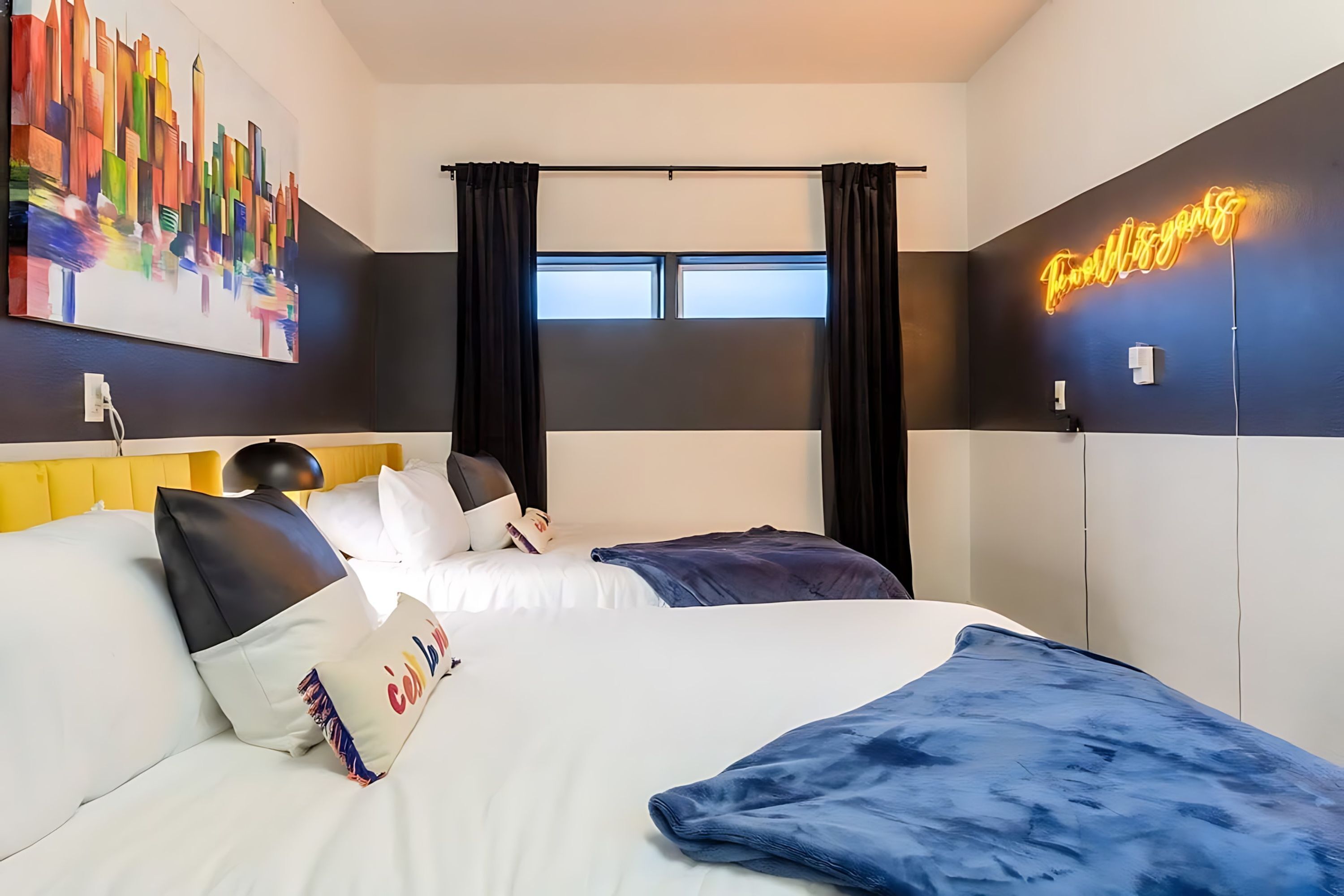Een moderne hotelkamer met een bed, een stadsgezicht schilderij, een neonbord en een raam met gordijnen.