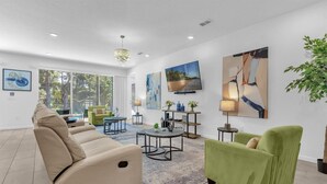 A modern living room with a beige sofa, a green armchair, and a coffee table. There are paintings on the walls and a large window providing natural light. - Upstay - Gator Getaway w Pool Hot Tub Theme Rooms (Kissimmee)