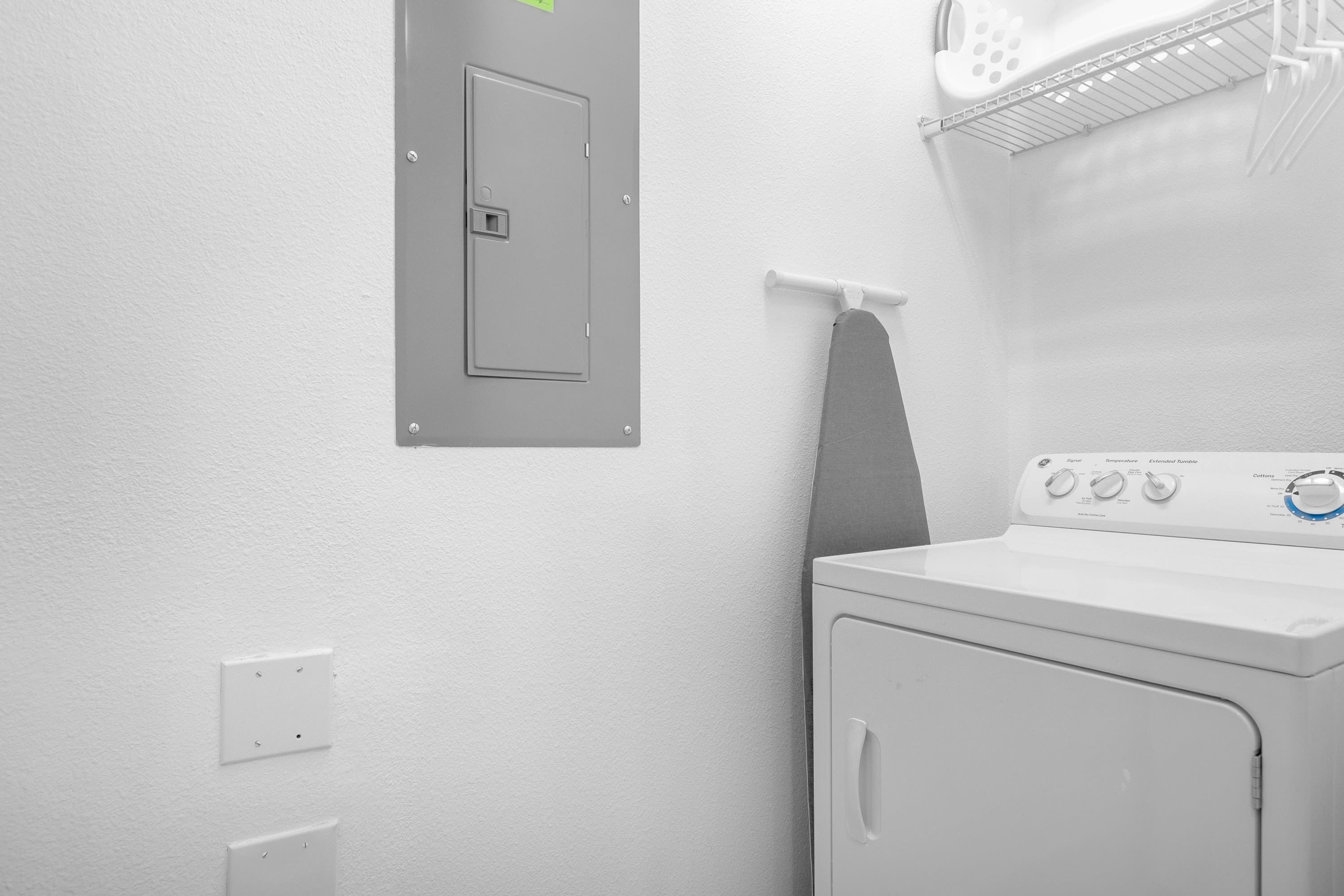 Un espace buanderie comprenant un lave-linge, une porte donnant sur les locaux techniques et une étagère en métal sur laquelle est posée une serviette blanche.
