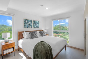 Ein Schlafzimmer mit einem großen Bett, zwei an der Wand befestigten Kunstwerken, einem Nachttisch mit Lampe und Blick ins Grüne durch ein Fenster.