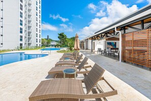 Ein Poolbereich mit Liegestühlen und Sonnenschirmen, ein Schwimmbecken und ein modernes Gebäude im Hintergrund.