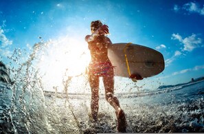 Eine Person mit Surfbrett steht im Wasser, im Hintergrund scheint die Sonne.