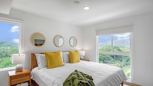 A bedroom with a large bed, two bedside tables, a nightstand, three round mirrors, and a window with a view of greenery. - Upstay - NEW Build - Viva Jaco Condo w Epic Views (Jacó)