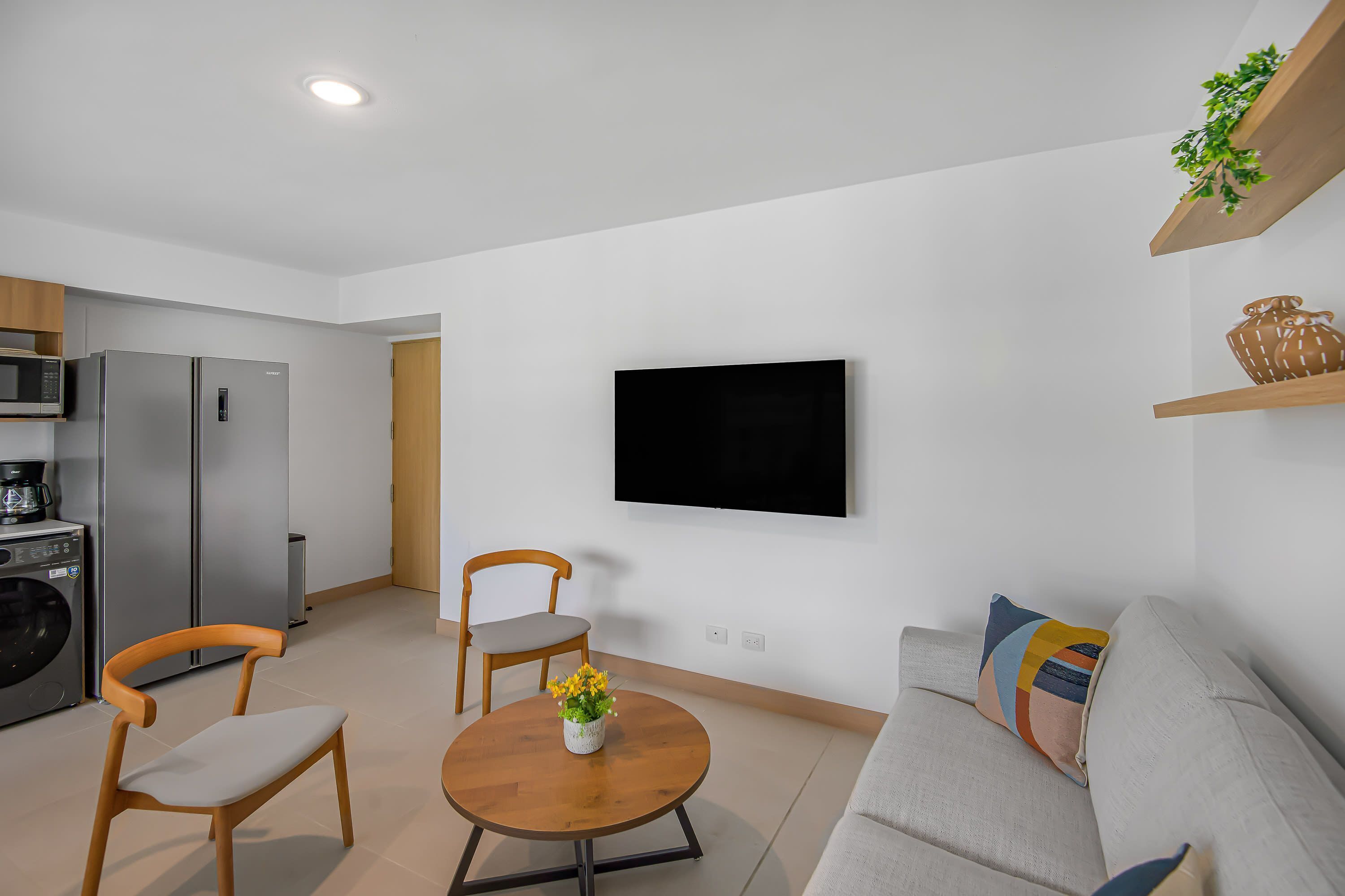 Ein modernes Wohnzimmer mit Sofa, zwei Sesseln, einem Couchtisch mit Blumen, einem flachbildfernseher an der Wand und einer Küchenzeile mit Geräte.