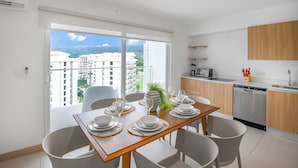 A modern kitchen with a dishwasher, a dining area with a table set for four, and a view of a cityscape through large windows. - Upstay - NEW Build - Viva Jaco Condo w Epic Views (Jacó)