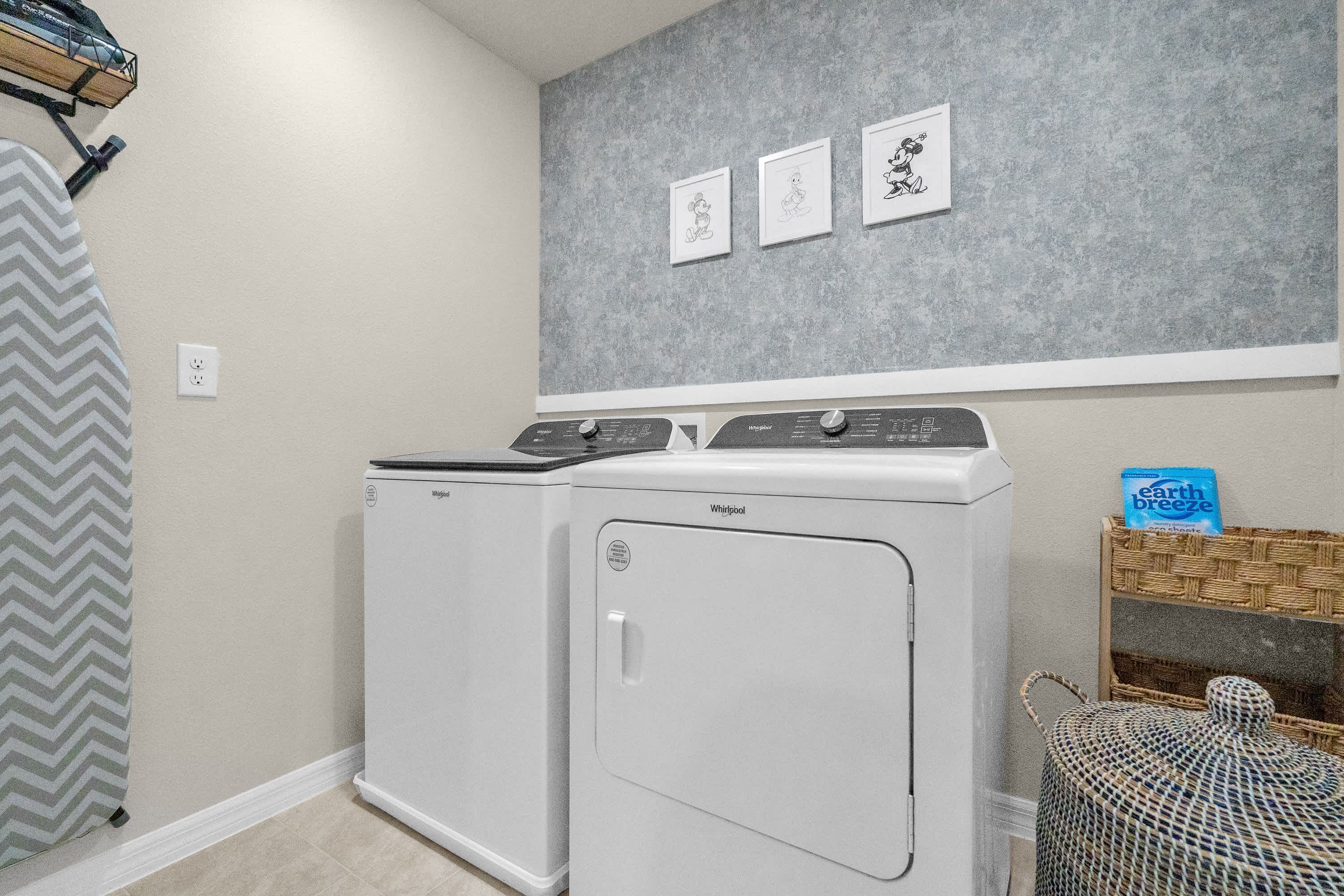 A laundry room with two white Whirlpool washers, a gray wall with three framed drawings, and a wicker basket with a blue bag on top.