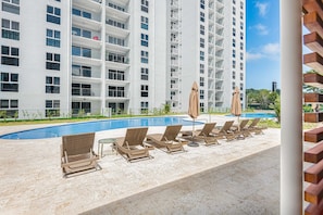 A poolside area with lounge chairs and umbrellas, a swimming pool, and a tall residential building in the background. - Upstay - Brand New Condo in Jaco 5mins to Beach (Jacó)