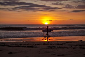 A sunset over the ocean with a person carrying a surfboard on a beach. - Upstay - Brand New Condo in Jaco 5mins to Beach (Jacó)