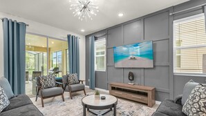Ein modernes Wohnzimmer mit großem Flachbildfernseher, grauen Sofas, einem Couchtisch und Blick ins Freie durch große Fenster.