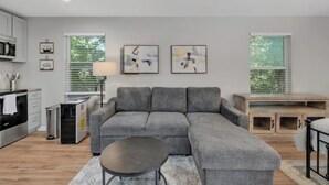 Ein modernes Wohnzimmer mit einer grauen Eckcouch, einem kleinen runden Tisch und einem Essbereich mit Holztisch und Bänken. Zwei Fenster mit Jalousien, eine Küche mit weißen Schränken und ein Deckenventilator sind ebenfalls vorhanden.