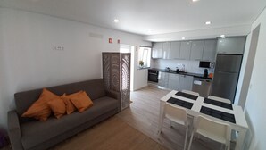 A modern living room with a grey sofa, orange cushions, a white dining table with chairs, and a kitchen area with grey cabinets and stainless steel appliances. - Nature, Naturally... (Cadaval)