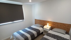 A bedroom with two beds, a window with blinds, a nightstand with a lamp, and wooden flooring. - Nature, Naturally... (Cadaval)