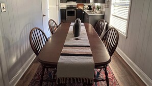 Un comedor con una mesa larga, sillas, un tapete y una cocina al fondo.