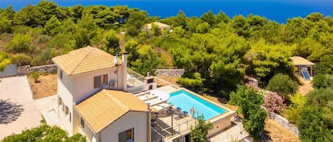 A two-story house with a swimming pool, surrounded by trees and a view of the sea.
