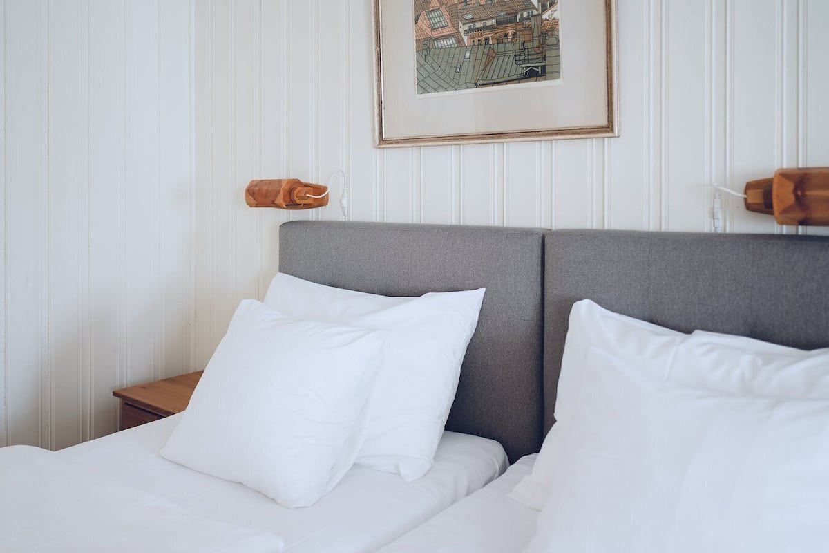 Una cama bien hecha con sábanas y almohadas blancas, un cabecero tapizado en gris y un cuadro en la pared.