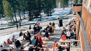 Evento al aire libre con personas sentadas y de pie sobre una terraza de madera, rodeado de terreno y árboles cubiertos de nieve.