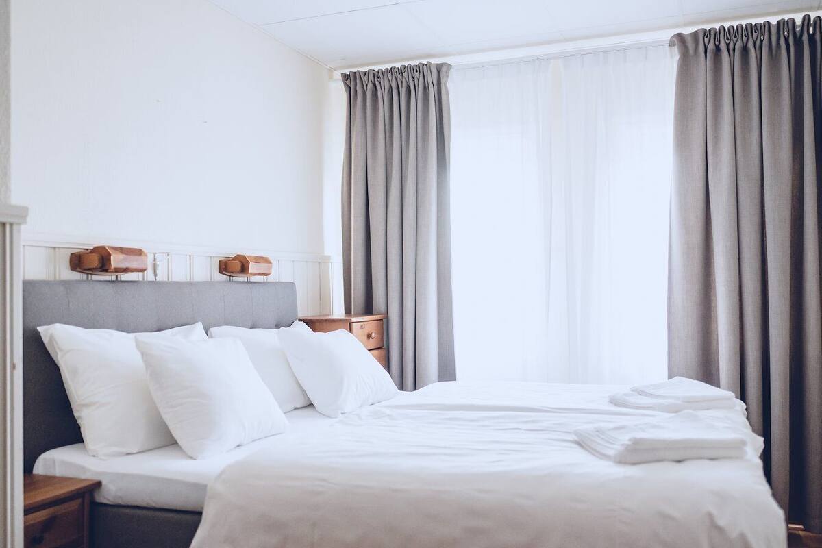 Una cama bien hecha con sábanas blancas, un cabecero gris y dos mesitas de noche con lámparas.