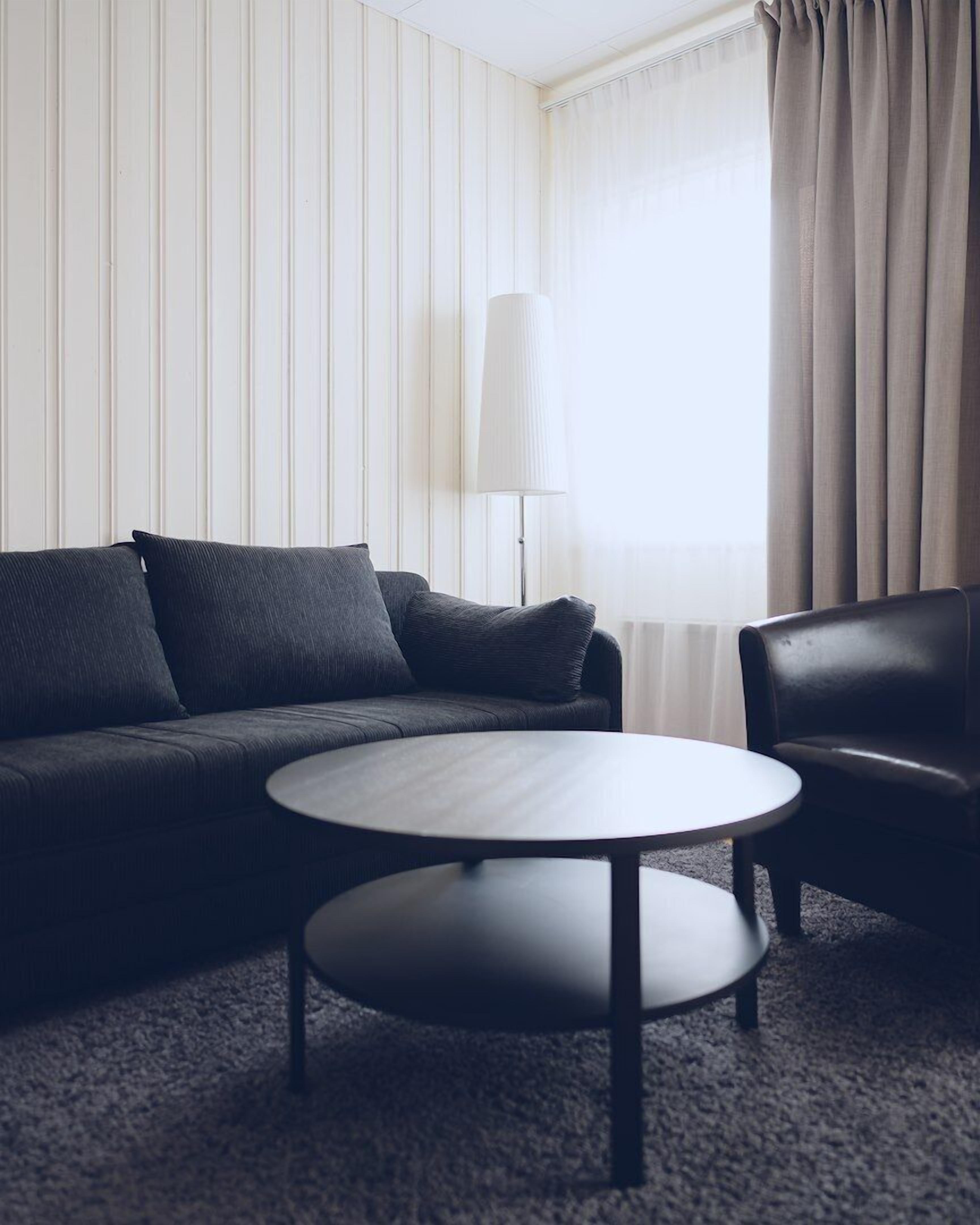 Een moderne hotelkamer met een donkergrijze bank, een ronde salontafel en een zwarte leren fauteuil.