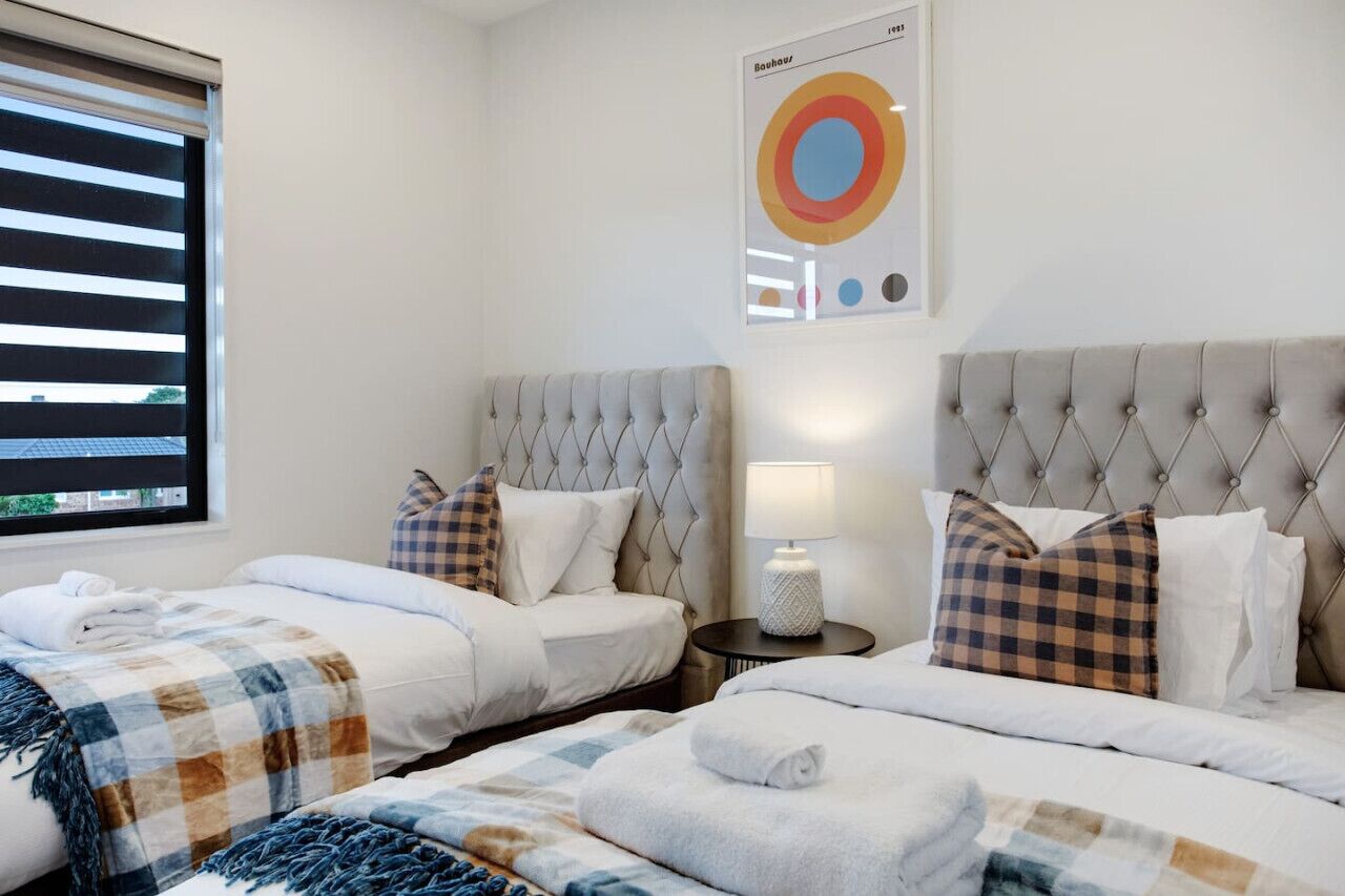 A hotel room with two beds, a window with blinds, a bedside table with a lamp, and a wall art piece.