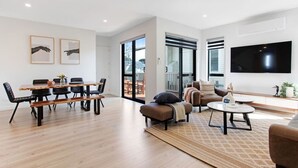 A modern living room with a wooden dining table, black chairs, a brown sofa, and a flat-screen TV mounted on the wall. - Central Oasis Townhouse with Parking and Air-con (Auckland Central Suburbs)