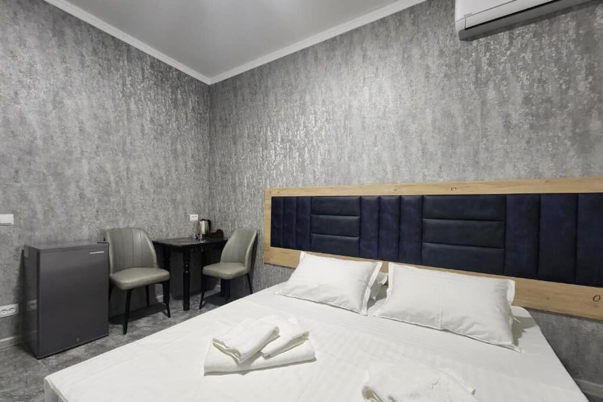 A hotel room with a bed, a desk, two chairs, a small refrigerator, and a wall-mounted air conditioner.