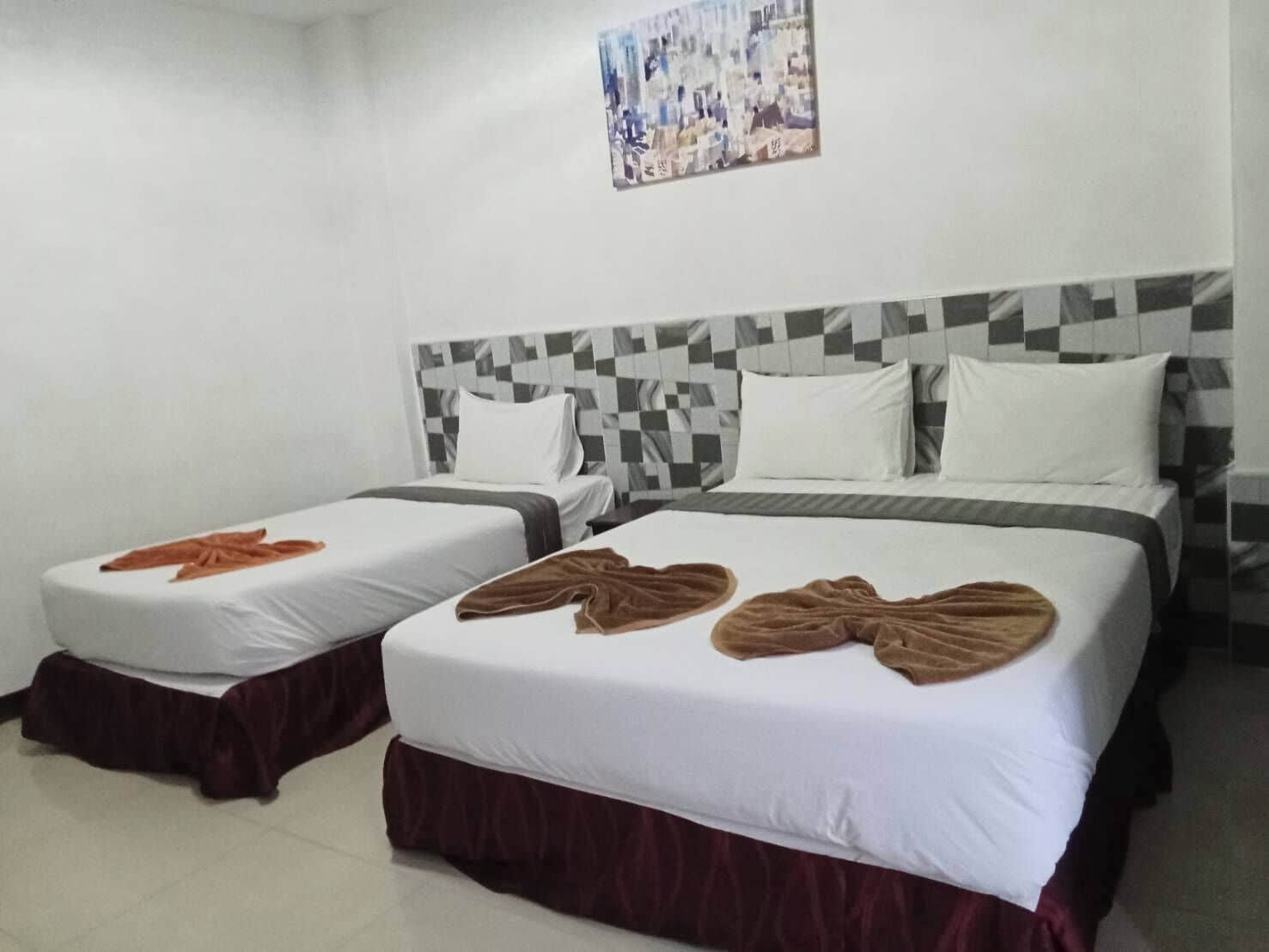 Ein Hotelzimmer mit zwei Betten, jeweils mit braunem Handtuch und weißen Laken. Der Kopfteil hat ein Schachbrettmuster, und über den Betten hängt ein Bild an der Wand.
