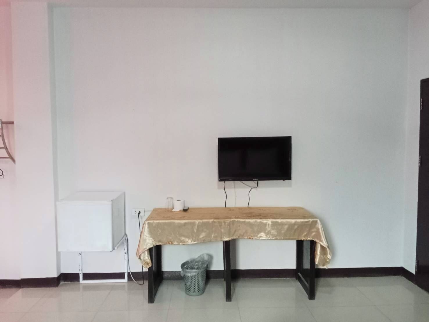 Una habitación con una televisión montada en la pared, una mesita con mantel, un cesto de basura y un armario blanco.