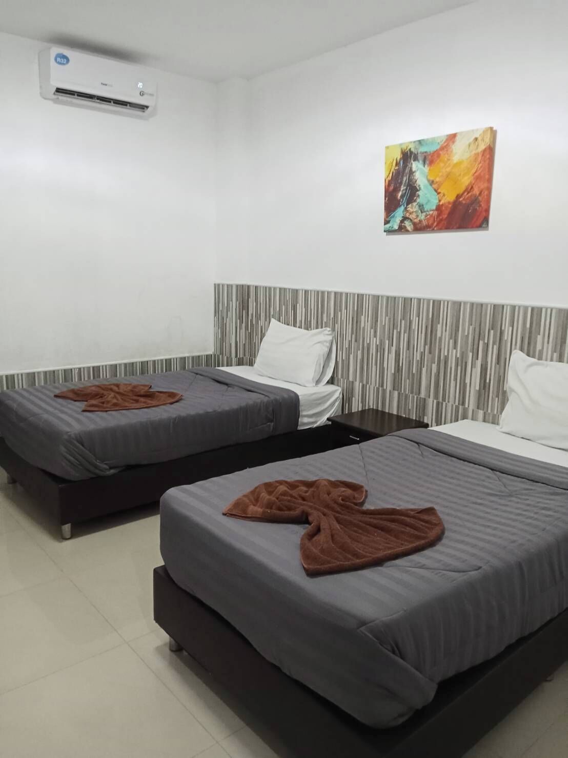 Habitación de hotel con dos camas individuales, cabecera a rayas grises y blancas, un cuadro abstracto de colores y una unidad de aire acondicionado instalada en el techo.
