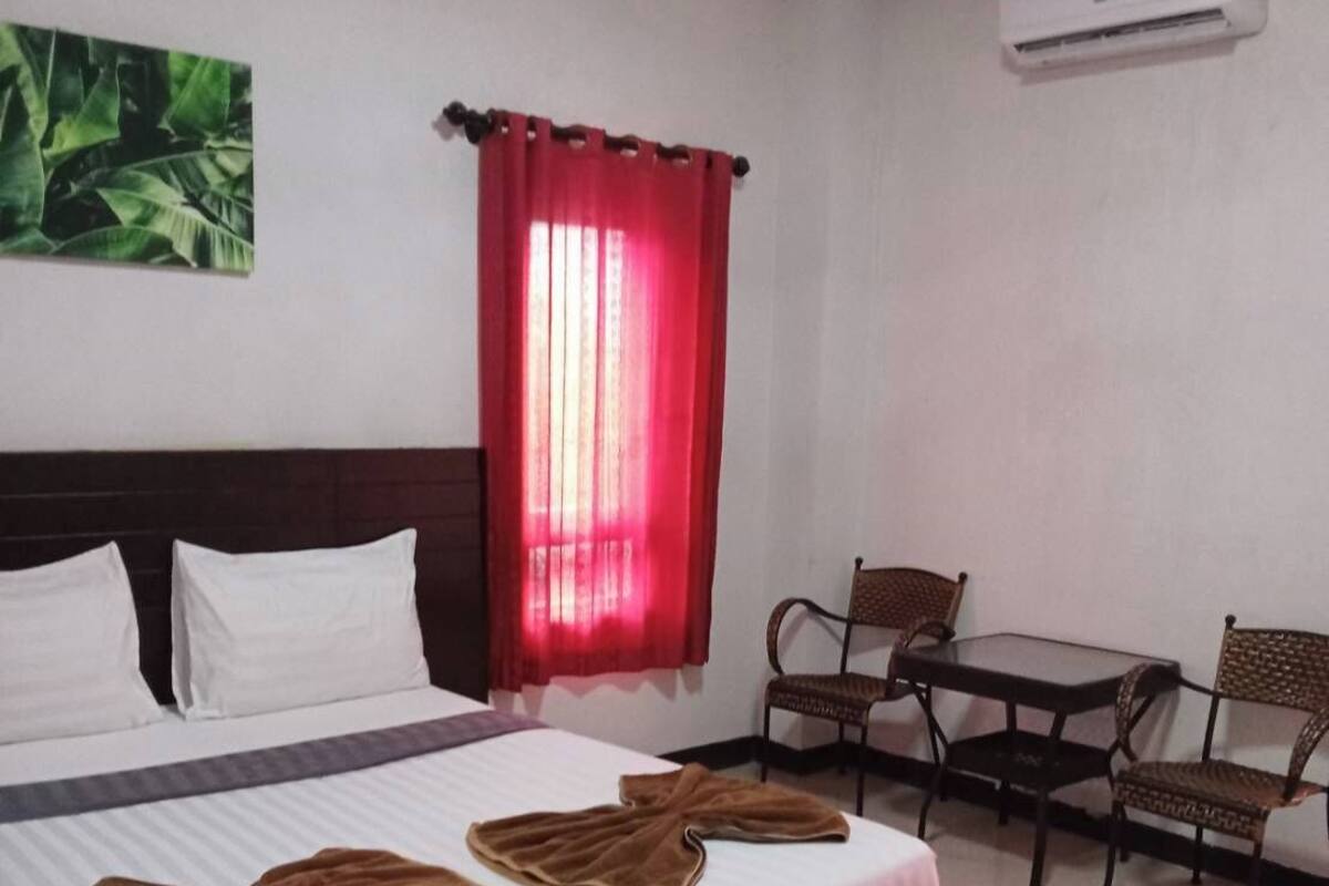 A hotel room with a bed, two chairs, a small table, a red curtain, and an air conditioning unit.