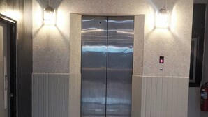 A modern elevator with metallic finish, flanked by wall-mounted lights, in a hallway with patterned wallpaper and tiled flooring. - PS Hotel Saraburi (Phra Phutthabat)